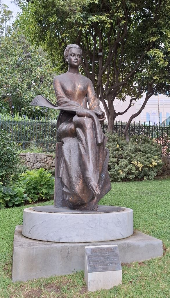 Bronzeskulptur einer sitzenden Frau in eleganter Pose, umgeben von grüner Landschaft und Bäumen.